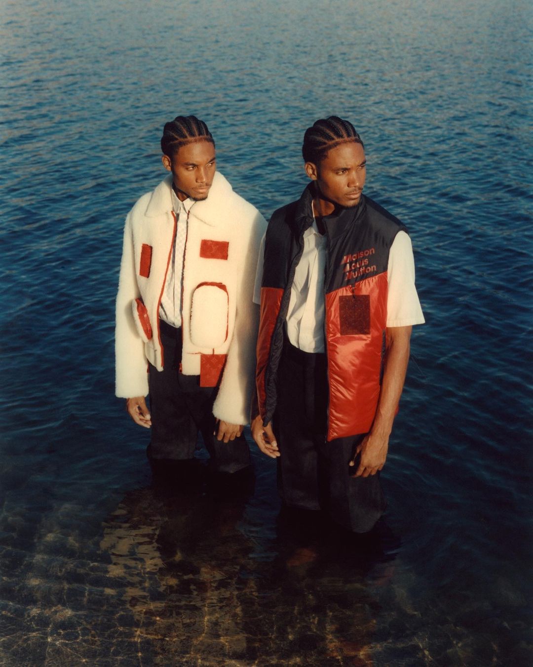 Two adult men wearing fashionable outerwear stand waist-deep in water with light reflecting on the surface in a medium shot.