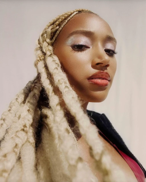 Close-up portrait of a woman with blonde braided hair, glitter eyeshadow, and glossy lips against a soft gradient background in studio lighting.