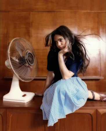 Young woman with long brown hair sits near a white electric fan. She wears a navy top and blue plaid skirt in indoor setting.