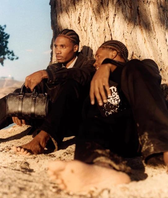 Two dark-skinned adult males with braided hair sit on a sandy surface beneath a tree, one holds a patterned handbag in an outdoor setting.
