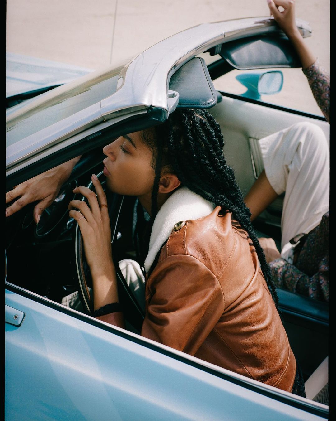 Two adults in a vintage blue convertible car. One adult with braided hair is in the driver's seat, wearing a brown jacket.