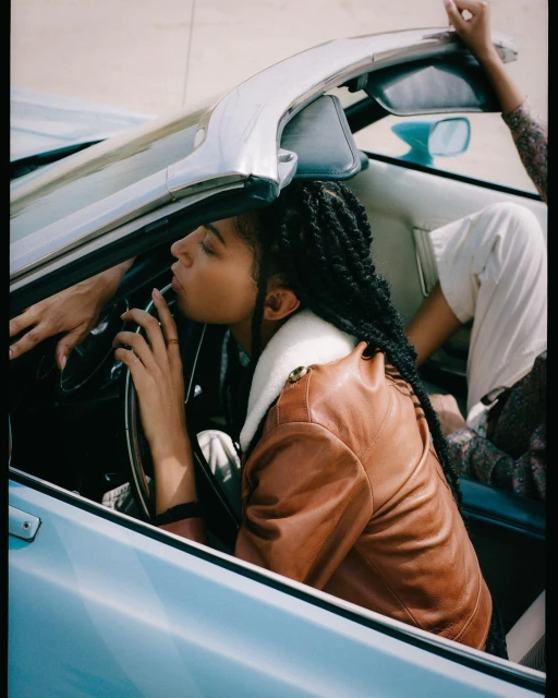 Two adults in a vintage blue convertible car. One adult with braided hair is in the driver's seat, wearing a brown jacket.
