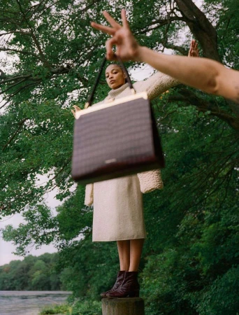 Model holding a brown structured handbag aloft outdoors against a green tree and water background by photographer Leeor Wild.