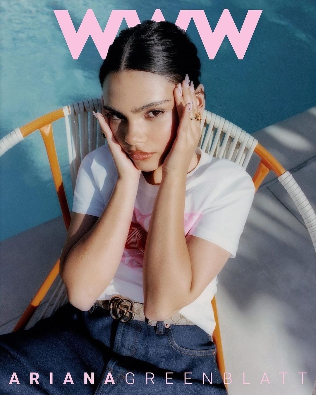 A woman with dark hair and blue jeans sits in a chair in front of a pool, with a pink W magazine logo on the image's top.