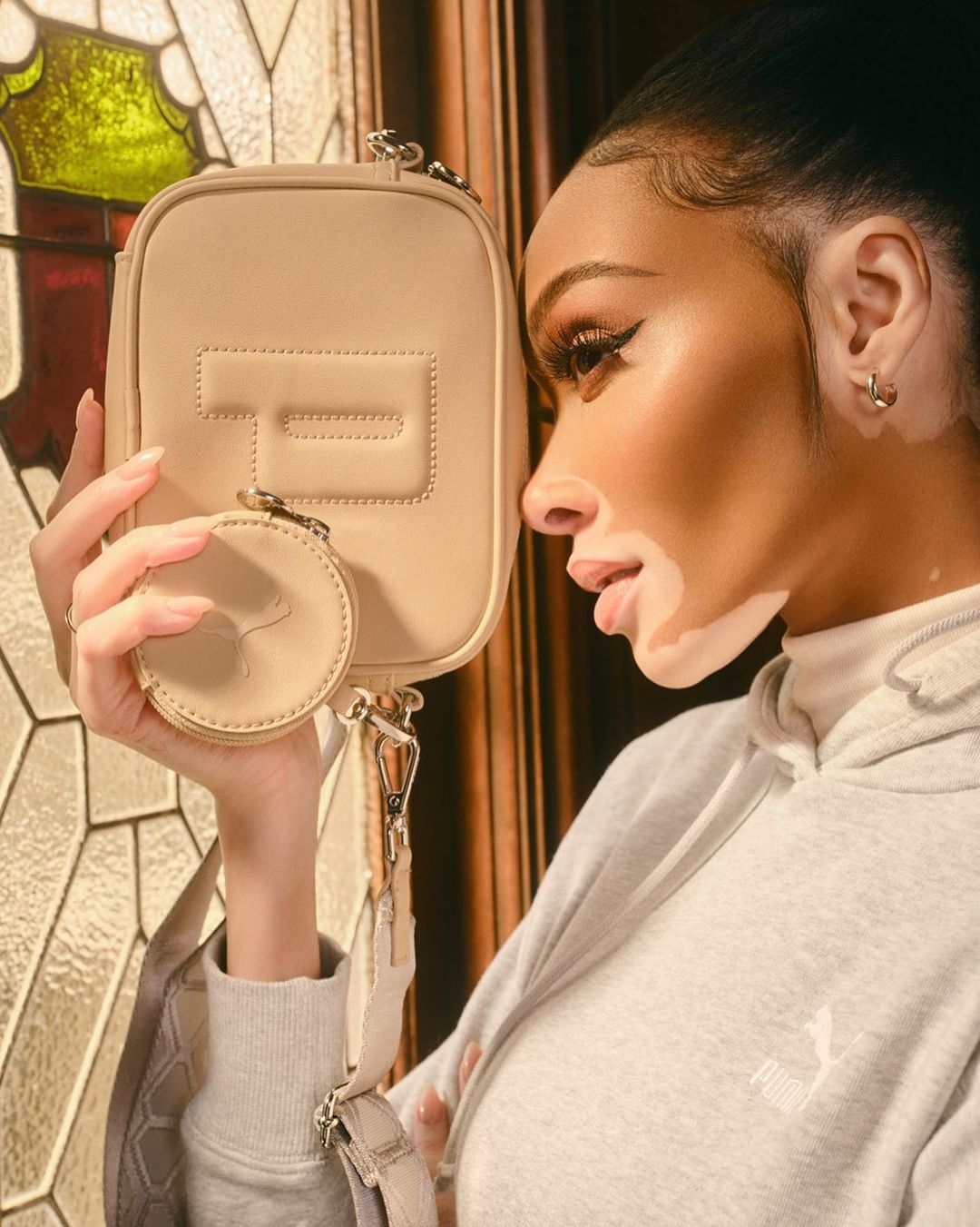 Adult woman poses in studio showcasing a small beige leather purse with round coin case and grey hoodie with Puma logo on the chest.