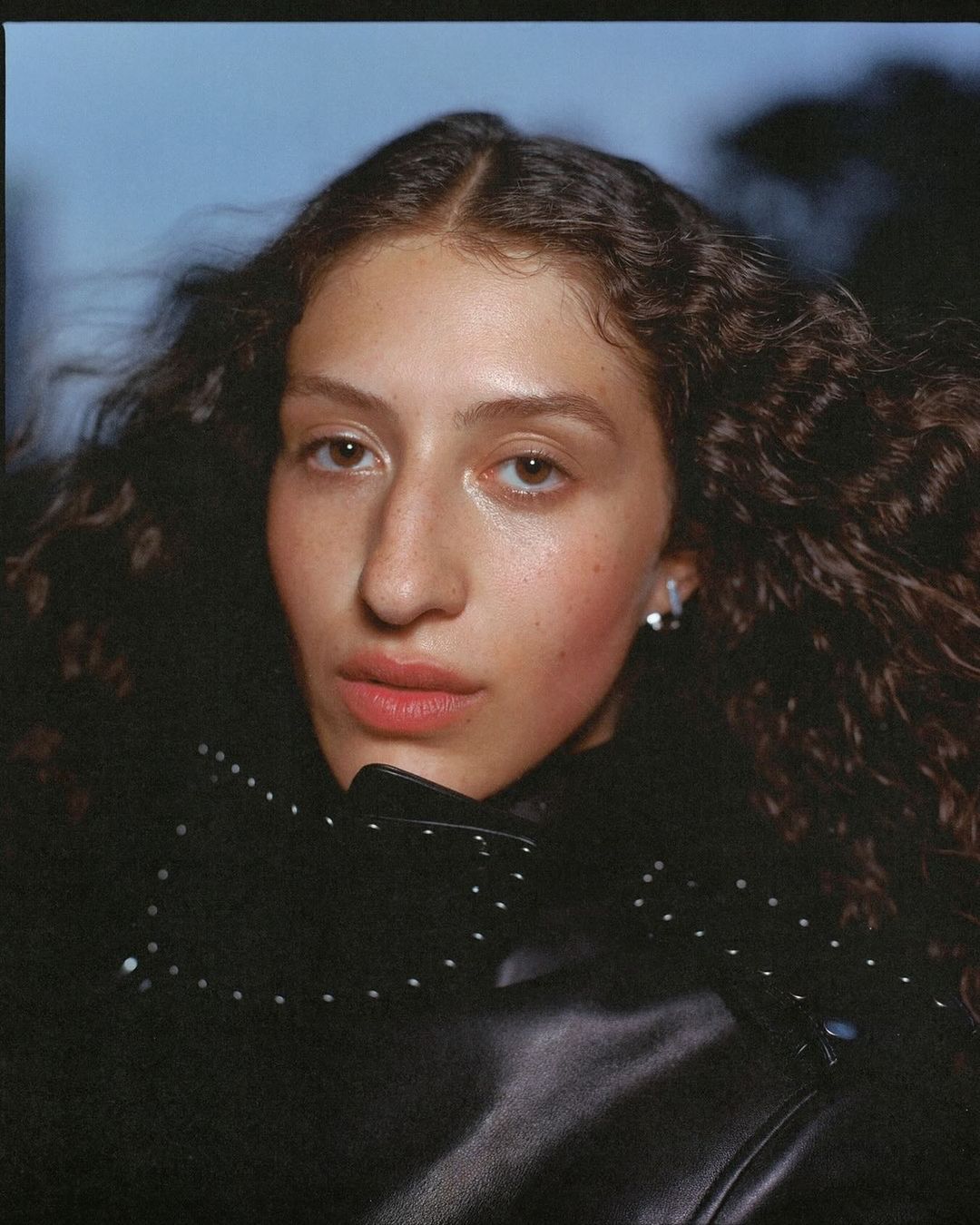 Portrait of a woman with curly hair wearing a black jacket, set against a blurred outdoor backdrop. Medium close-up, eye-level angle.