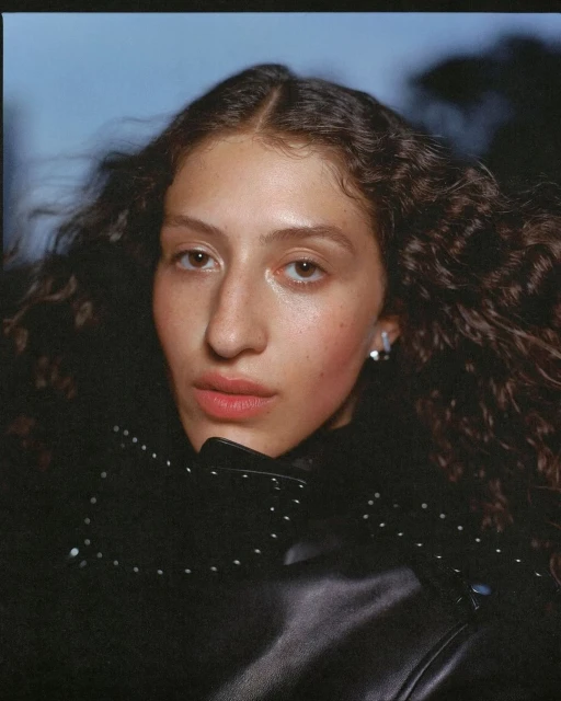 Portrait of a woman with curly hair wearing a black jacket, set against a blurred outdoor backdrop. Medium close-up, eye-level angle.