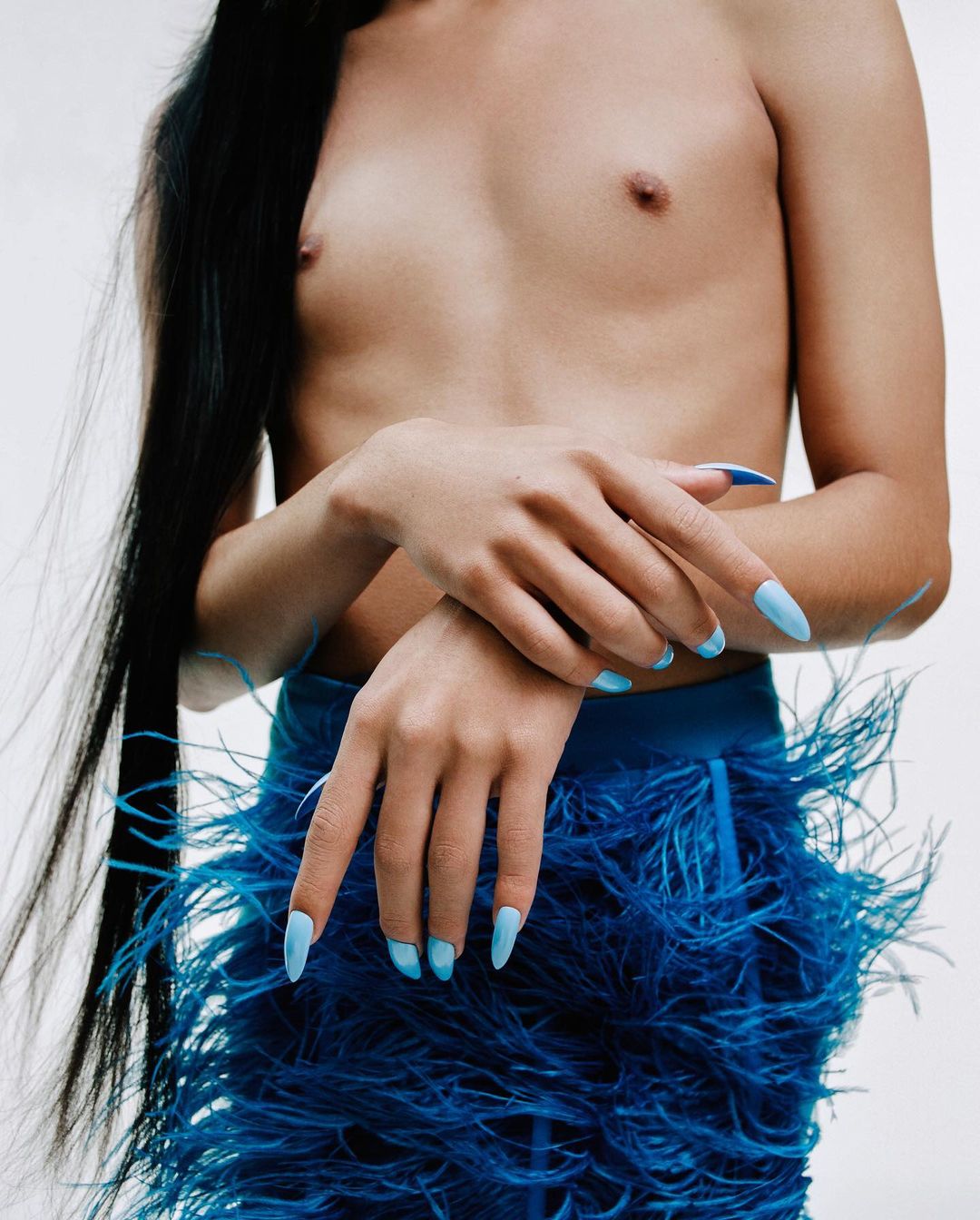 Model poses with long dark hair, blue feather skirt and light blue stiletto nails against a white background in a studio shot.