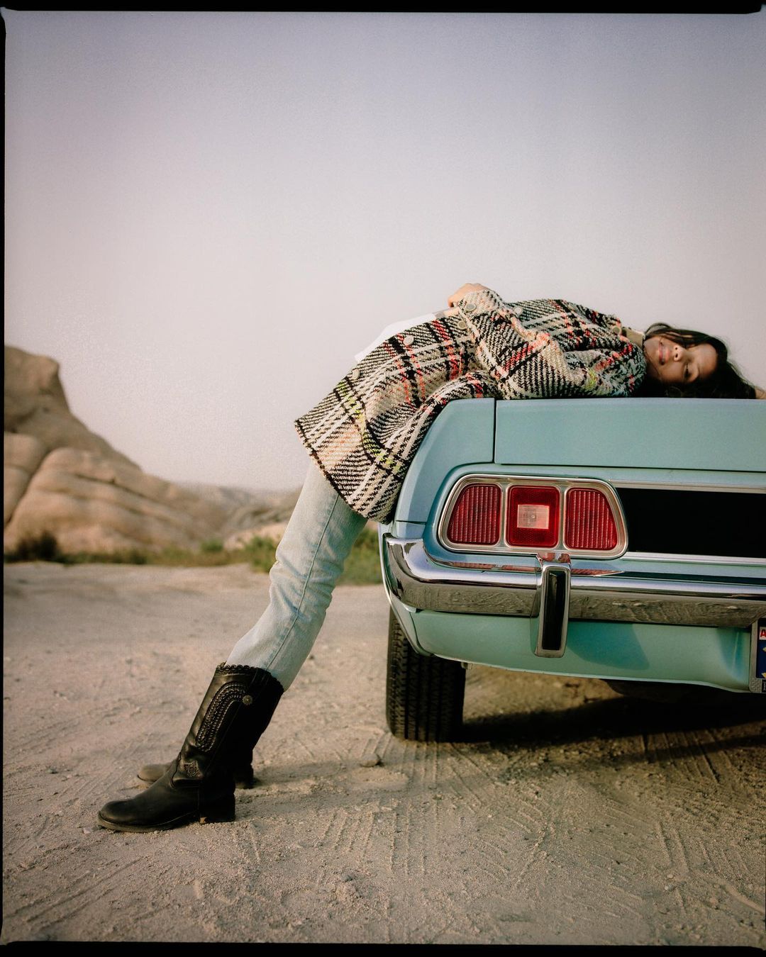 Woman in plaid coat reclines on the trunk of a vintage turquoise car against a desert backdrop, wearing cowboy boots.