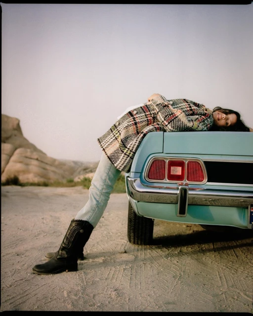 Woman in plaid coat reclines on the trunk of a vintage turquoise car against a desert backdrop, wearing cowboy boots.