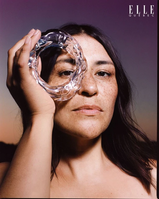 Portrait of a freckled woman holding a clear, wavy ring to her eye, creating a distorted view, with Elle Québec logo.