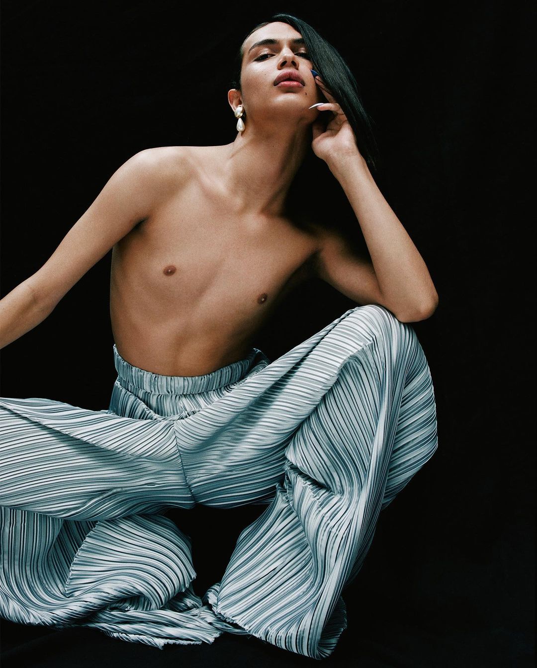Transgender model posing with light blue pleated pants on black background, medium shot. Fashion, beauty, and gender identity concept.
