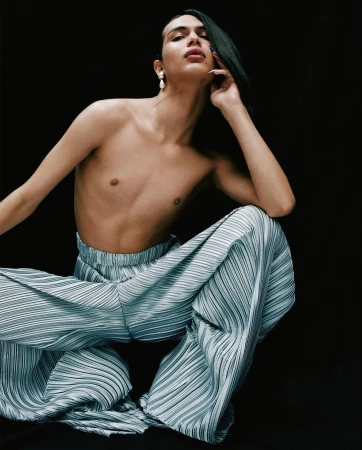 Transgender model posing with light blue pleated pants on black background, medium shot. Fashion, beauty, and gender identity concept.