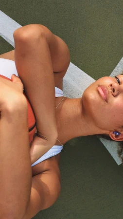 Woman in white tank top laying on green sports court hugging basketball with eyes closed, sunlit, vertical orientation.
