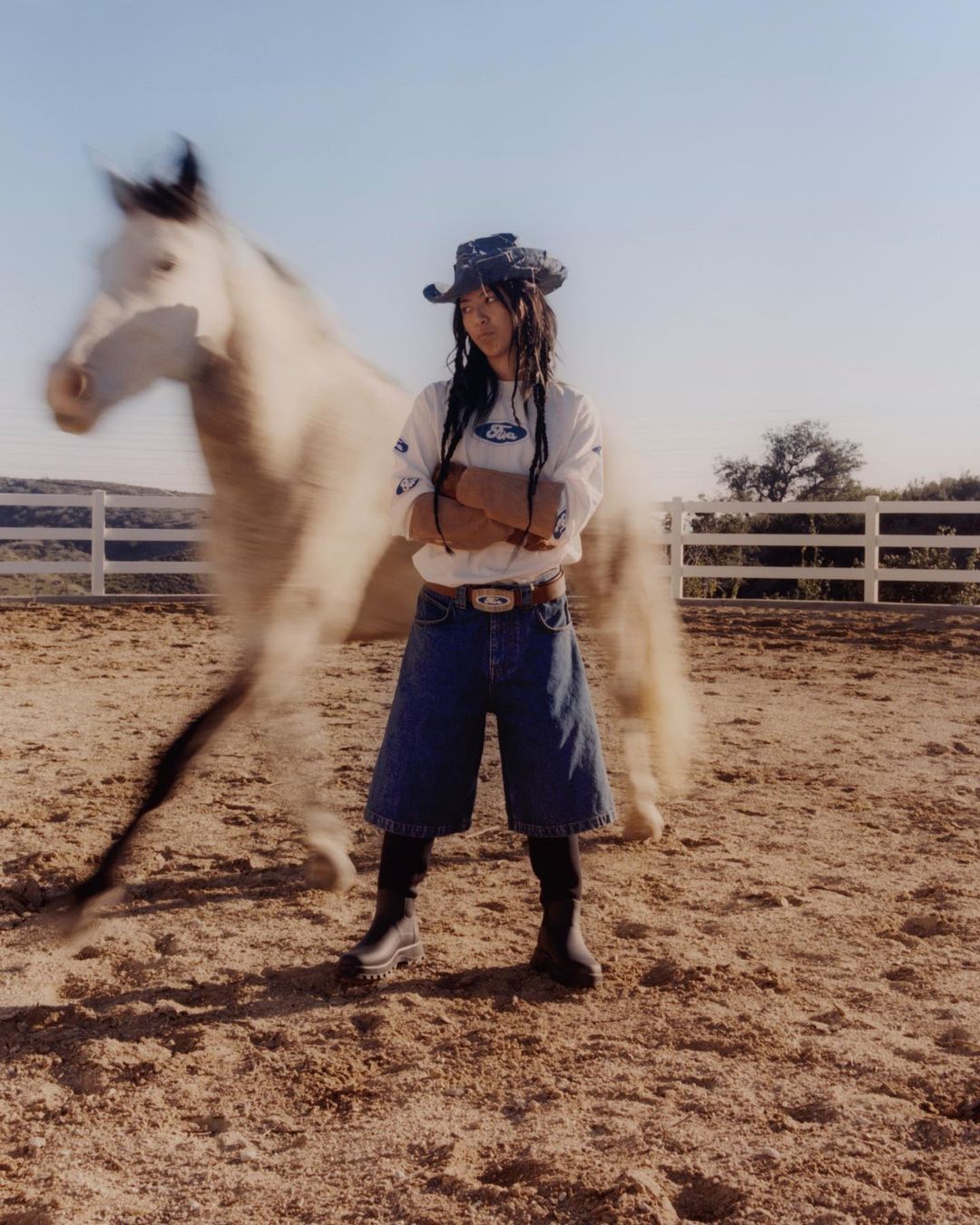 Woman in cowboy hat with arms crossed stands near a blurred white horse in an outdoor dirt paddock with a wooden fence.