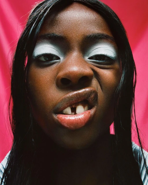 Close-up of a young person making a face at the camera against a pink backdrop, featuring bold white eyeshadow and a gap-toothed smile.