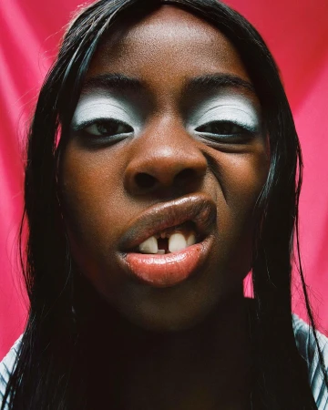 Close-up of a young person making a face at the camera against a pink backdrop, featuring bold white eyeshadow and a gap-toothed smile.