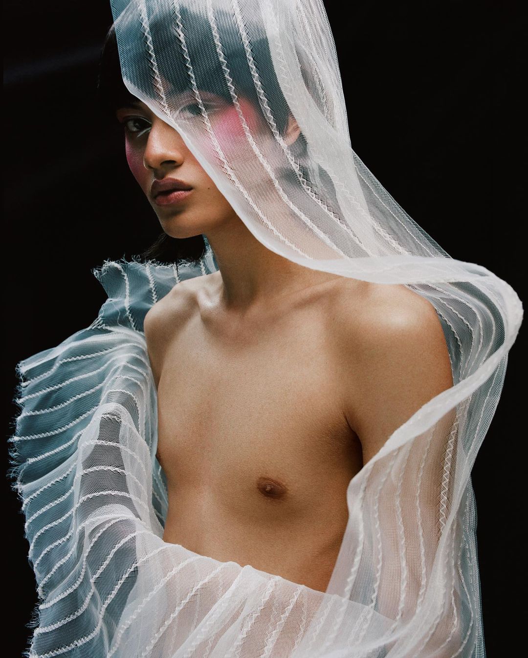 Portrait of a shirtless person with pale skin and sheer white fabric draped over their body, standing in a dark studio setting.