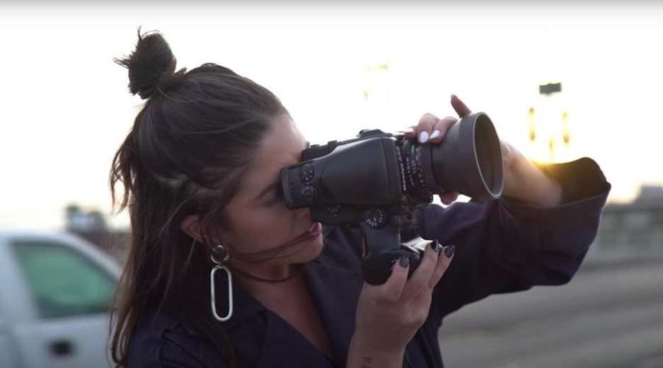 Adult female photographer aiming professional camera outdoors in daytime, wearing a dark blue coat and silver hoop earrings.