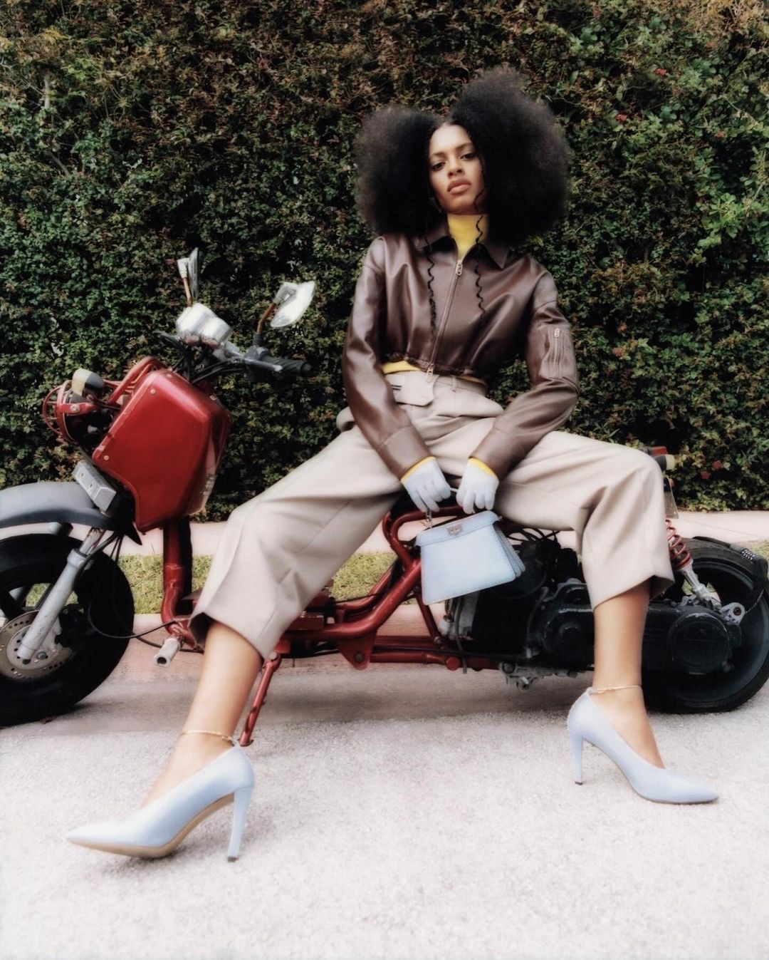 Stylish woman in brown jacket and cream pants poses on a red mini motorcycle outside with lush green hedge background, holding a light blue purse.