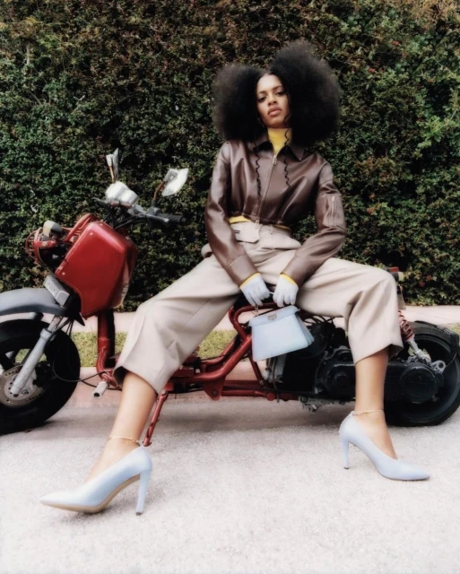 Stylish woman in brown jacket and cream pants poses on a red mini motorcycle outside with lush green hedge background, holding a light blue purse.