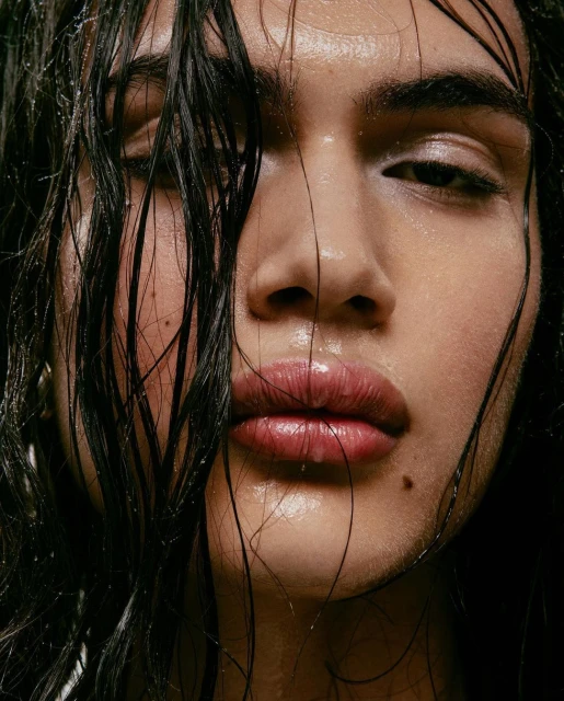 Close-up portrait of a person with dark, wet hair, capturing their face with shiny, moisturized skin and a focused gaze, against a dark background.