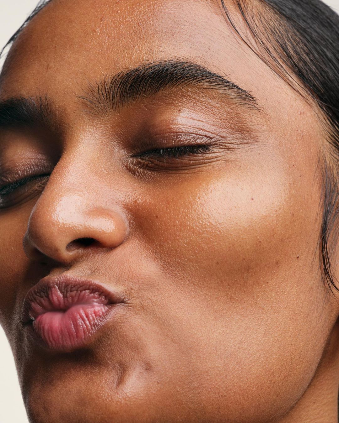 Close-up of an adult woman with brown skin and closed eyes, puckering her lips in a kiss face expression, against a neutral background.