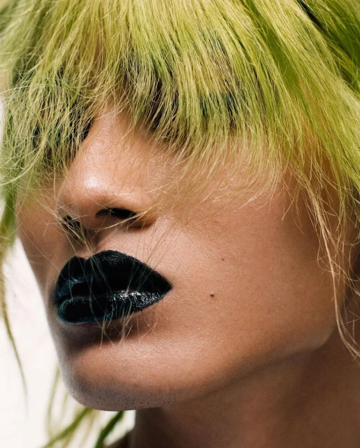 Close-up portrait of a person with green-yellow hair and black lipstick against a white background, showcasing bold beauty aesthetics.