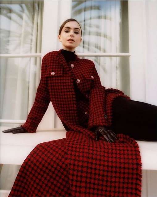 Elegant adult female poses in a red houndstooth coat and black gloves while seated in front of a window with diffused natural light.