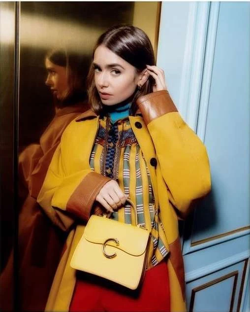 A stylish woman in a yellow coat holds a matching purse, with a blurred reflection visible in a modern indoor setting.