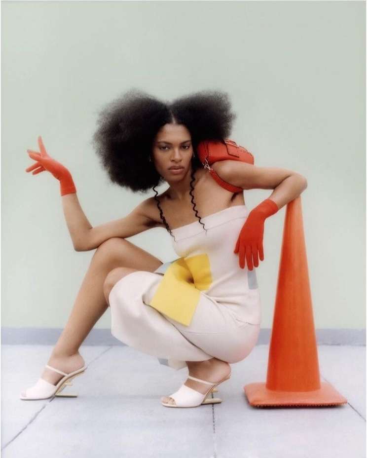 Adult female fashion model poses in a studio with a red handbag, safety cone, cream jumpsuit and red gloves against a light green backdrop.