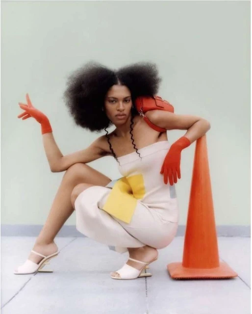 Adult female fashion model poses in a studio with a red handbag, safety cone, cream jumpsuit and red gloves against a light green backdrop.