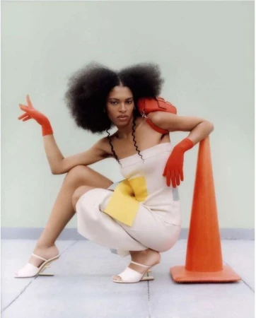 Adult female fashion model poses in a studio with a red handbag, safety cone, cream jumpsuit and red gloves against a light green backdrop.