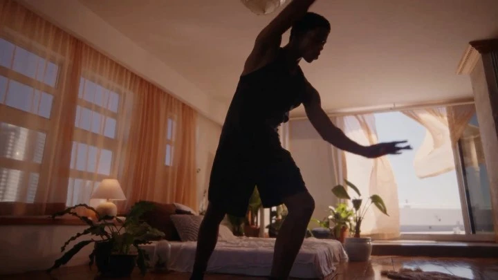 A Black man dancing inside a sunlit bedroom with sheer orange curtains and a low bed, captured by Leeor Wild.