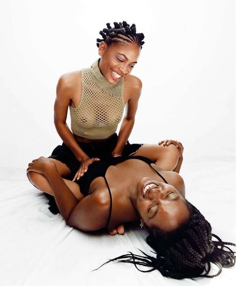 Two Black women laugh together in a light studio. One sits cross-legged while the other lies across her lap, wearing casual tops and hairstyles.