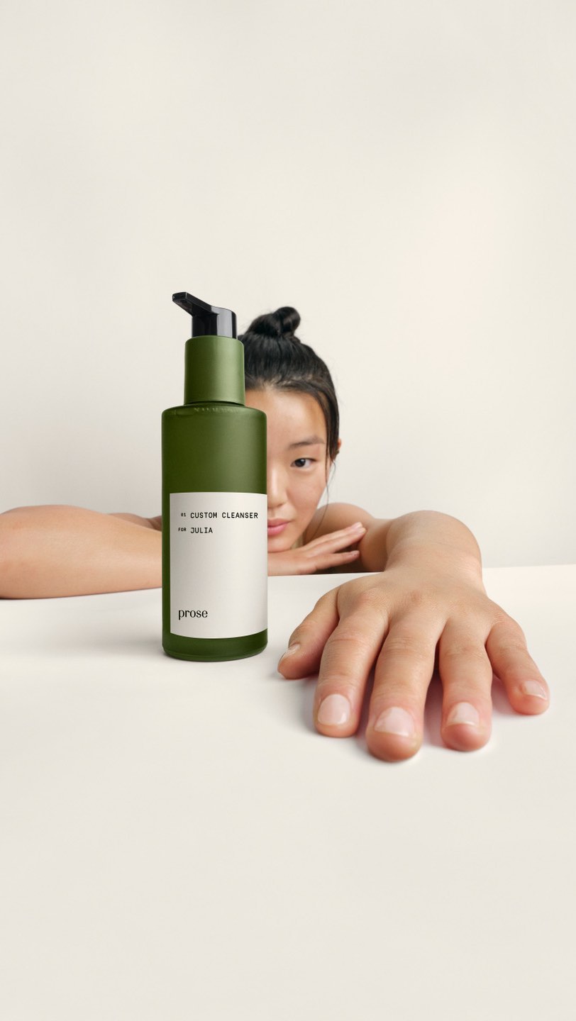 Olive green bottle of cleanser with pump dispenser, resting on a white surface with a woman's face and hand visible in the frame.
