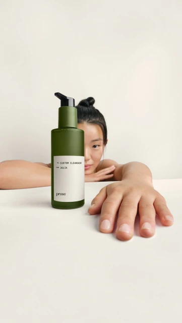 Olive green bottle of cleanser with pump dispenser, resting on a white surface with a woman's face and hand visible in the frame.