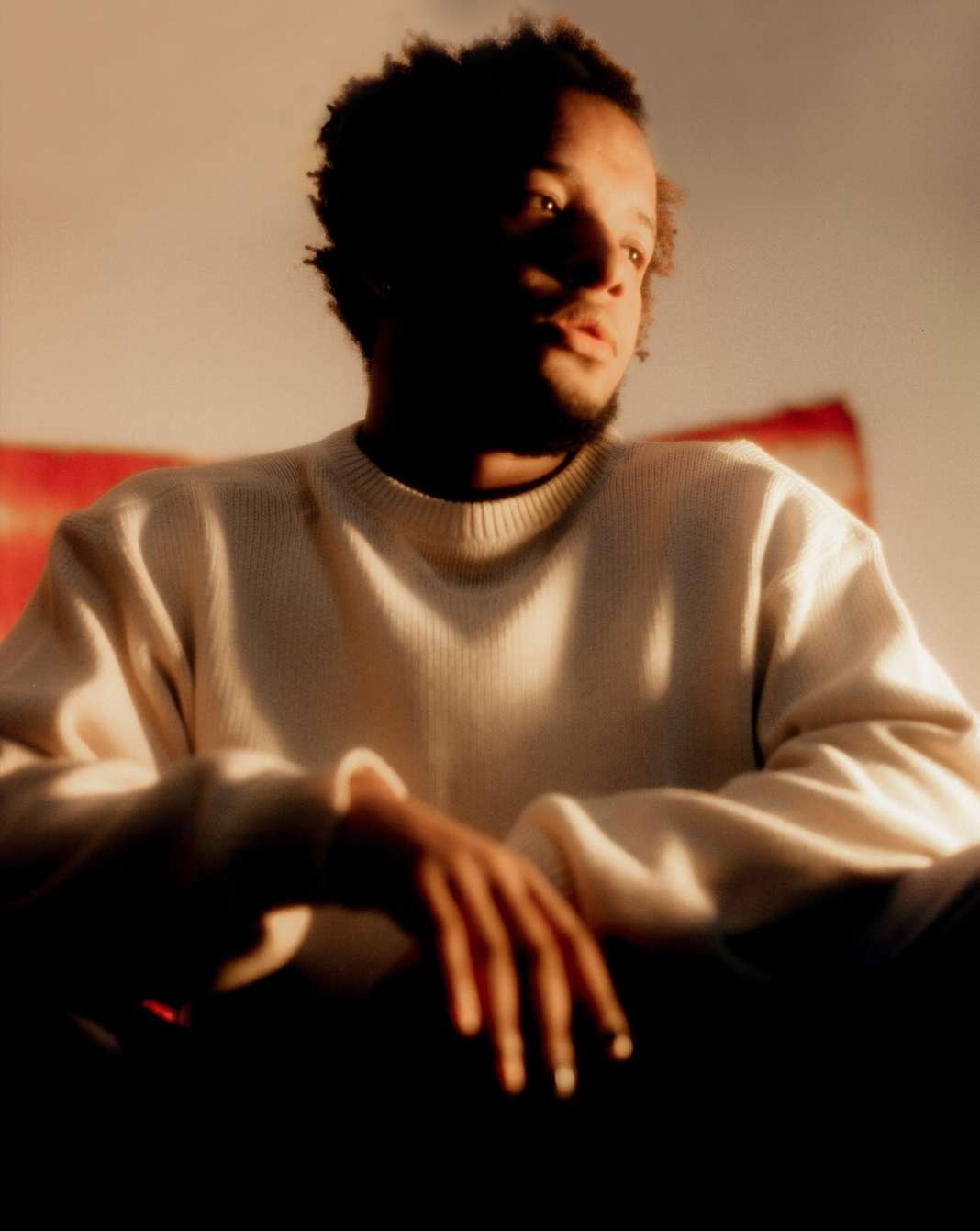 Portrait of an adult Black male in a cream sweater, looking left with hands clasped, under warm lighting against a muted backdrop.