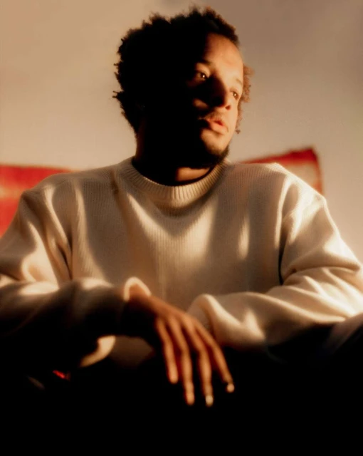 Portrait of an adult Black male in a cream sweater, looking left with hands clasped, under warm lighting against a muted backdrop.