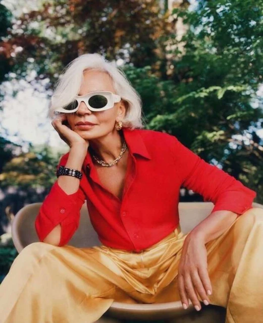 Older woman with short white hair wearing sunglasses, a red shirt and gold pants sits in a chair outside surrounded by green foliage.