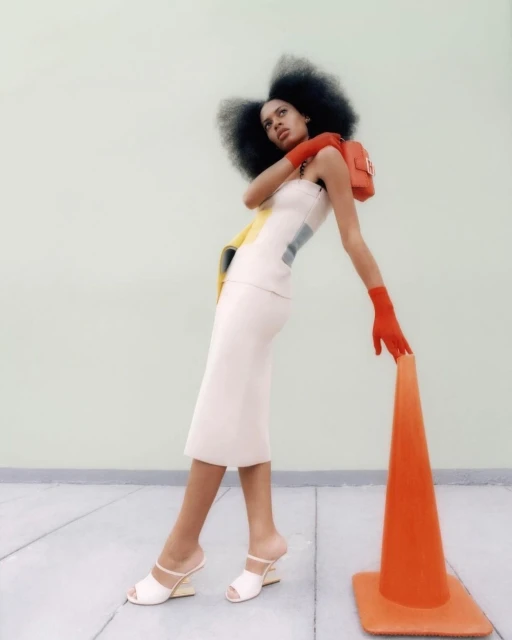 Fashion shot of a woman in white dress and red gloves leaning on an orange traffic cone with a purse on her shoulder.