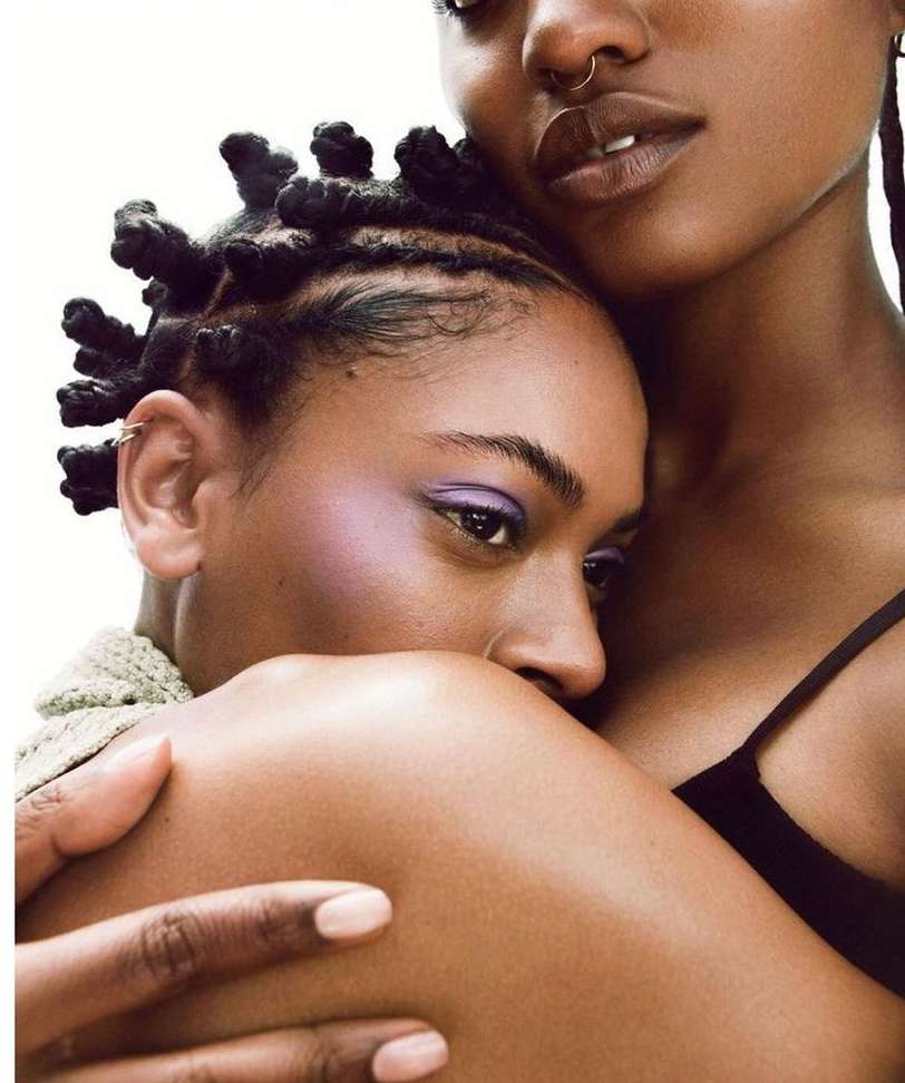 Close-up portrait of two Black women embracing, one with braided knot hair and the other with a nose ring, against a white background.