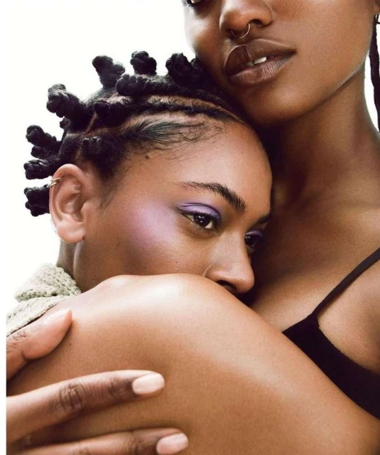 Close-up portrait of two Black women embracing, one with braided knot hair and the other with a nose ring, against a white background.
