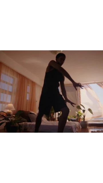 An adult stands in a bedroom with arms extended, soft light entering through the window, and indoor plants adding a natural touch.