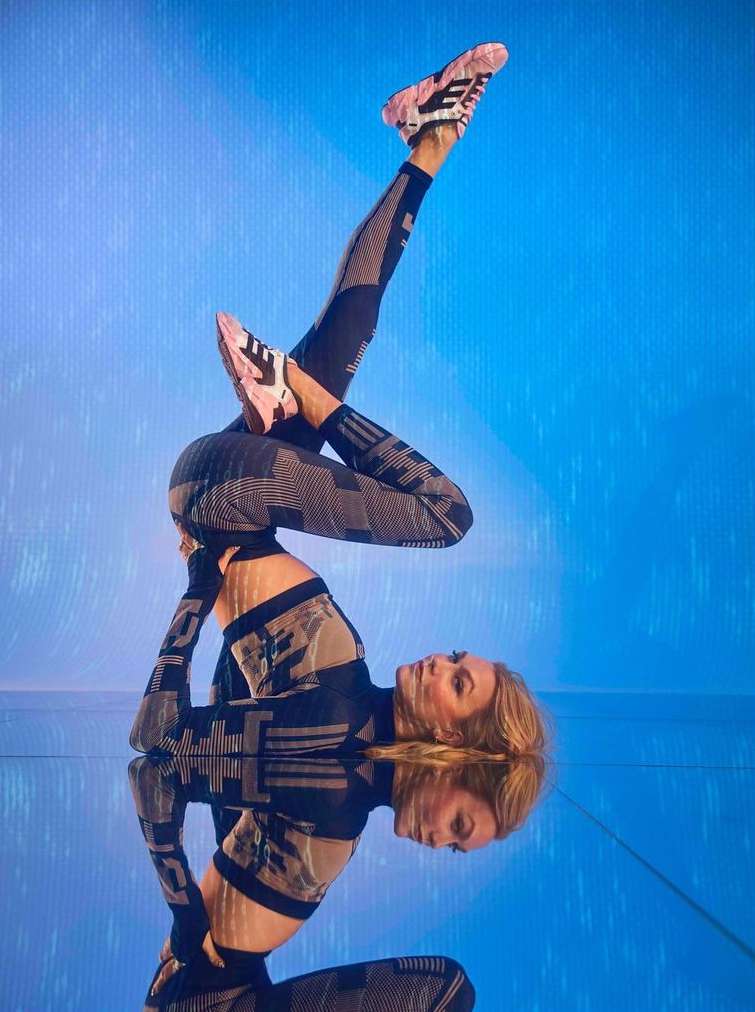 Blonde woman in athletic wear balances upside down on a reflective surface with her feet pointed up in pink and black sneakers.
