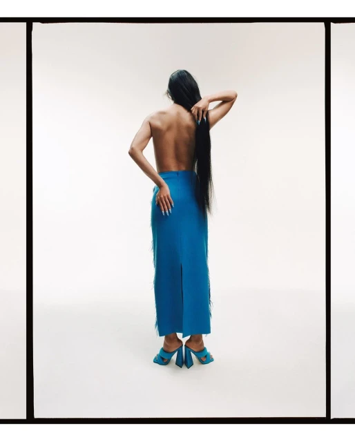 Back view of adult woman with long hair, bare upper body, blue fringe skirt, and blue mules in a studio setting with a white background.