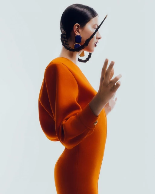Profile view of a woman in an orange dress with braided hair, square earrings, and a neutral expression against a white backdrop.