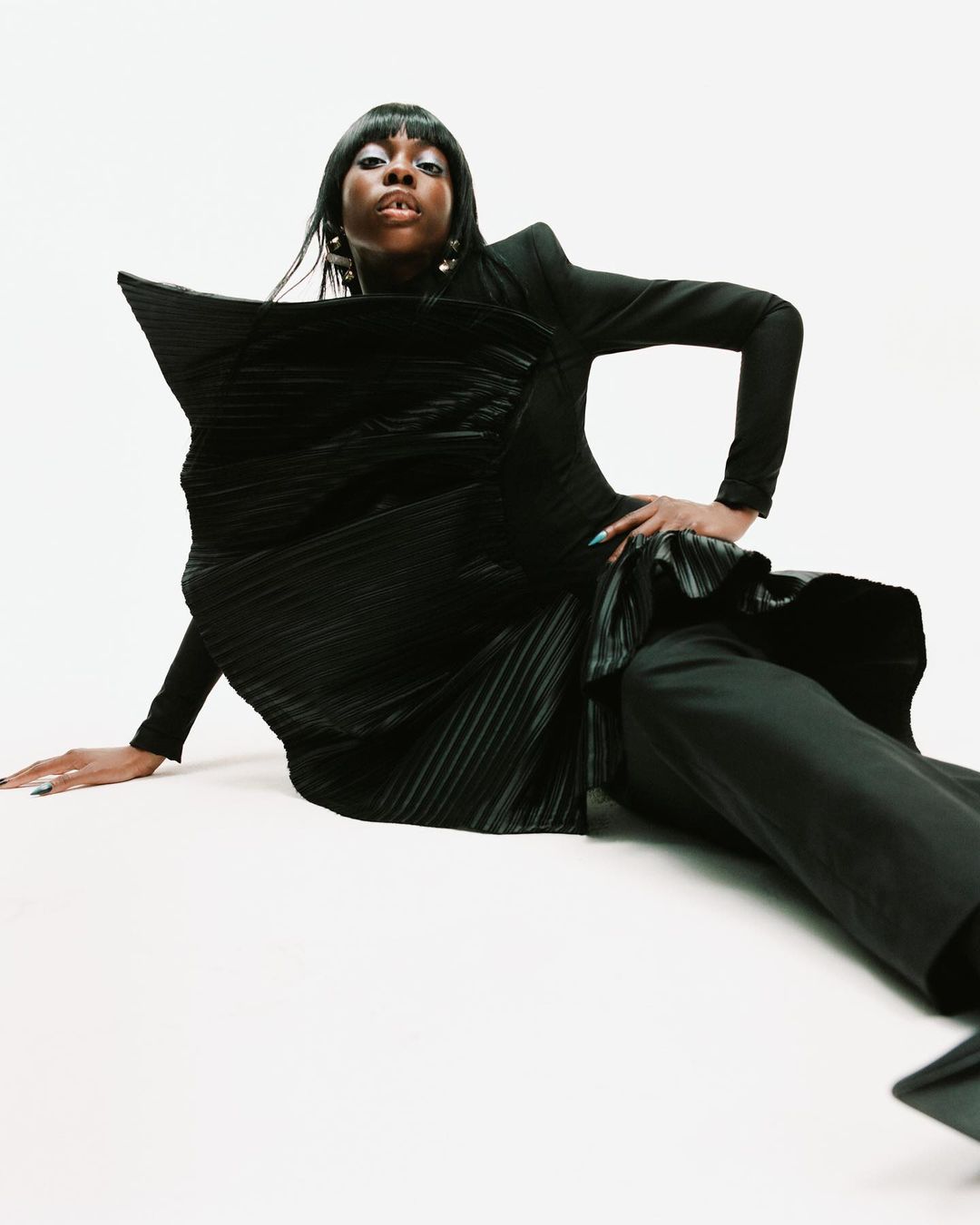 Portrait of a Black woman in a black sculptural dress, sitting on a white background. Her dark hair frames her face, and she wears gold earrings.
