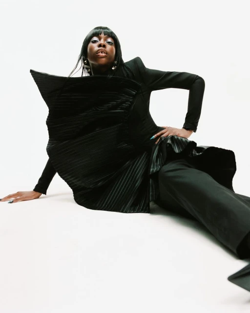 Portrait of a Black woman in a black sculptural dress, sitting on a white background. Her dark hair frames her face, and she wears gold earrings.
