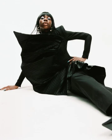 Portrait of a Black woman in a black sculptural dress, sitting on a white background. Her dark hair frames her face, and she wears gold earrings.